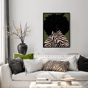 Op de afbeelding: Een ingelijste kunstdruk met een vrouw met een grote afro en kleding met zebra-strepen tegen een groene achtergrond. Het kunstwerk hangt boven een witte bank met decoratieve kussens en een zwarte vaas met takken.