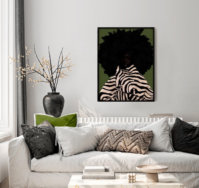 Op de afbeelding: Een ingelijste kunstdruk met een vrouw met een grote afro en kleding met zebra-strepen tegen een groene achtergrond. Het kunstwerk hangt boven een witte bank met decoratieve kussens en een zwarte vaas met takken.