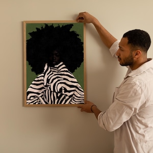 Op de afbeelding: Een ingelijste kunstdruk met een portret van een persoon met een grote, donkere afro en een jas met zebraprint. De achtergrond is een gedempt groen. Het kunstwerk wordt aan een lichtgekleurde muur gehangen.