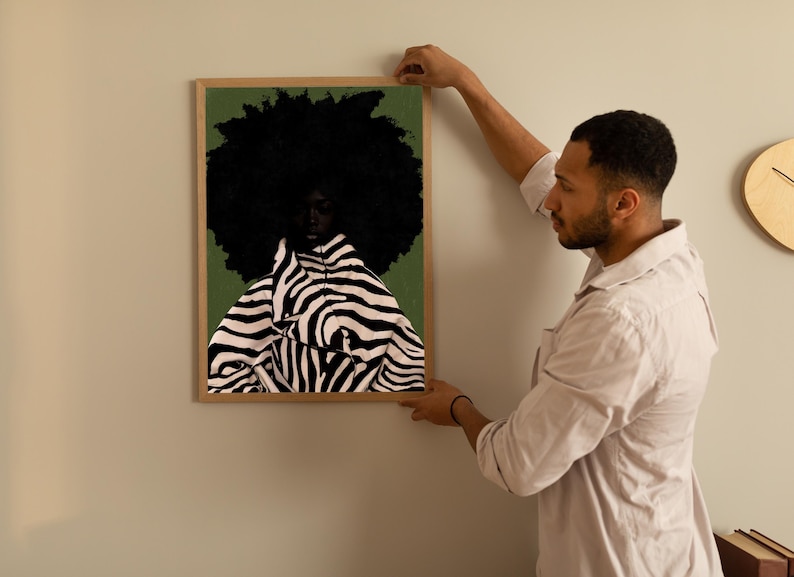 Op de afbeelding: Een ingelijste kunstdruk met een portret van een persoon met een grote, donkere afro en een jas met zebraprint. De achtergrond is een gedempt groen. Het kunstwerk wordt aan een lichtgekleurde muur gehangen.