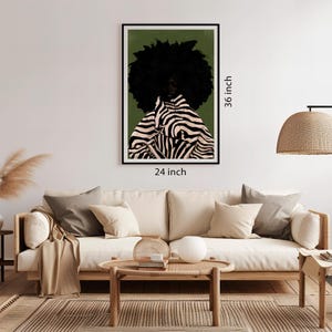 Op de afbeelding: Een ingelijste kunstdruk met een persoon met een grote zwarte afro en een jas met zebraprint. Het kunstwerk is 61 cm breed en 91 cm hoog. De achtergrond is een gedempt groen en de lijst is zwart. De kunst wordt boven een beige bank weergegeven.