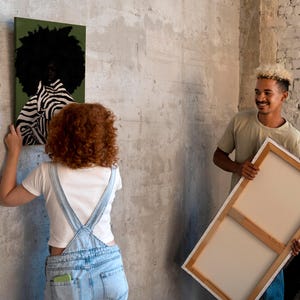 Op de afbeelding: Twee mensen hangen canvas kunst op. E&eacute;n canvas toont een persoon met een afro en zebra-gestreepte kleding tegen een groene achtergrond. Het andere canvas is leeg. De kunst wordt opgehangen aan een getextureerde, grijze muur.