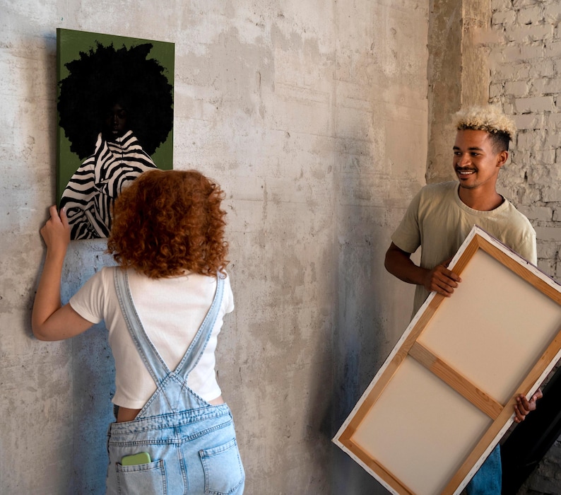 Op de afbeelding: Twee mensen hangen canvas kunst op. E&eacute;n canvas toont een persoon met een afro en zebra-gestreepte kleding tegen een groene achtergrond. Het andere canvas is leeg. De kunst wordt opgehangen aan een getextureerde, grijze muur.
