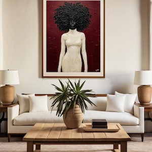 May include: A framed artwork depicts a woman with a large, textured black afro and a white turtleneck dress against a deep red background. The artwork hangs above a white sofa with white pillows, a wooden coffee table, and two lamps.