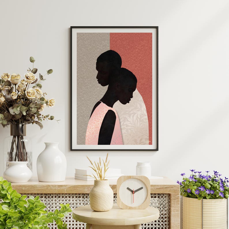 May include: A framed wall art print featuring a stylised illustration of two dark-skinned figures in profile against a split background of textured grey and terracotta red. The figures wear a light pink garment with a subtle leaf pattern.