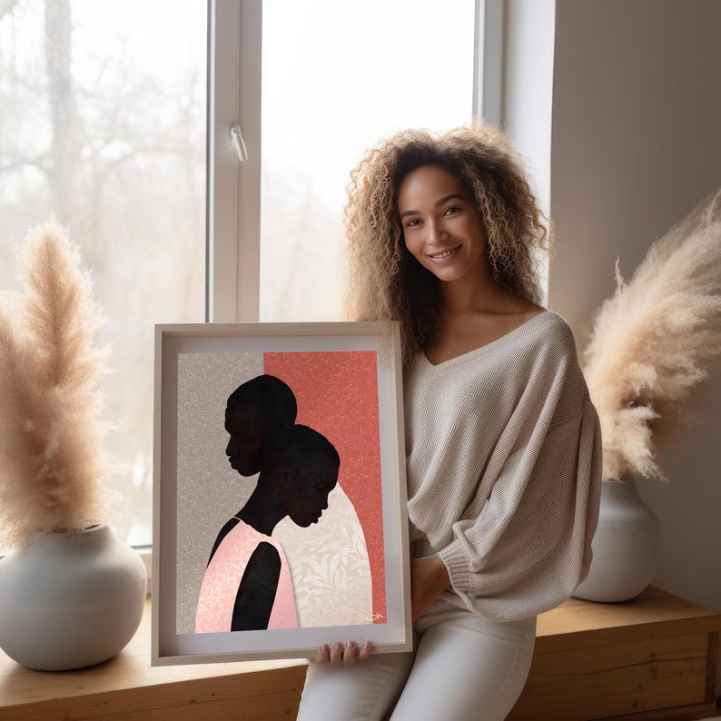 May include: A framed art print featuring a stylised illustration of two dark-skinned figures in profile against a split background of textured light grey and coral pink, with one figure wearing a light pink garment and a subtle leaf pattern.