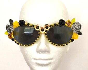 May include: Yellow and black sunglasses with floral embellishments. The sunglasses have black lenses and are decorated with yellow and black flowers, gold beads, and black and white patterned accents.