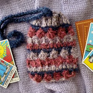 Puede incluir: Una bolsa de crochet de color morado, rosa y azul, sobre una tela de punto morado. La bolsa contiene cartas de tarot, incluyendo las cartas "Rey de Copas" y "Estrella".