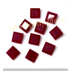 May include: A collection of nine square-cut, deep red gemstones. Each gem has a lighter, square-shaped center, creating a framed effect. The gems are scattered on a white surface, showcasing their color and cut. The image is well-lit, highlighting the facets.