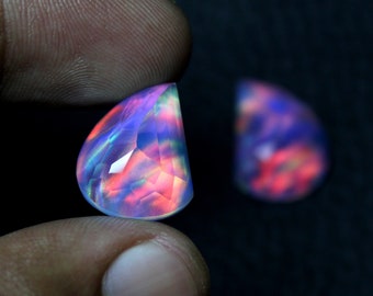 May include: A faceted, semi-circular opal gemstone, held between fingers, displays a vibrant play of color including blues, pinks, and greens. A second, out-of-focus opal is visible in the background. The gemstone is cut to maximize light refraction.