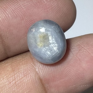 May include: A pale blue, oval gemstone with an uneven surface. The gem has a milky appearance with a touch of yellow. The stone is held between fingers, showing its size and shape. The gem is likely a sapphire.