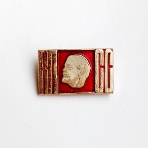May include: A gold-colored pin with a red background and a profile of a man's face. The pin has the text "CCCP" in gold letters.