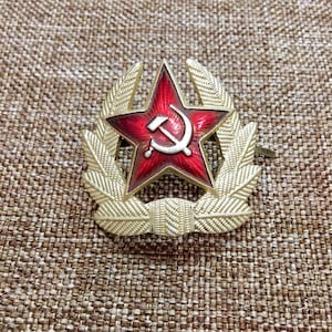 Può includere: Una stella di metallo dorato con un centro in smalto rosso che presenta un martello e una falce. La stella è circondata da due corone di alloro in metallo dorato.