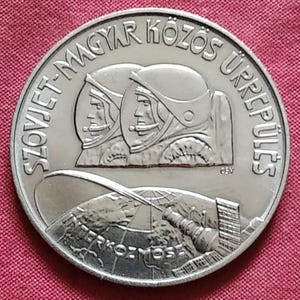 May include: A silver coin featuring two astronauts in helmets, with text in a foreign language around the edge. The coin also depicts a satellite and a structure with the word "INTERKOZMOSZ" at the bottom.