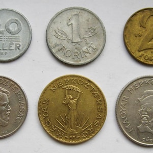 May include: Six Hungarian coins of varying designs and denominations. The coins are silver and gold in colour, with visible text and dates. The coins are arranged on a light background.