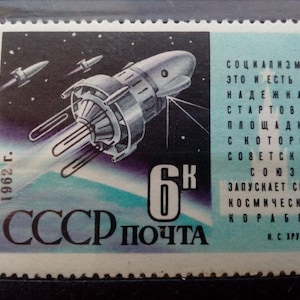 Può includere: Un francobollo sovietico vintage che presenta un'illustrazione in bianco e nero di un satellite nello spazio. Il francobollo è blu e bianco con il testo "1962 г. 6K СССР ПОЧТА" e una citazione in russo sul lancio della sua navicella spaziale da parte dell'Unione Sovietica.