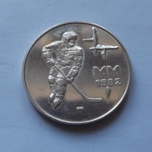 May include: A silver coin featuring a hockey player in action, with a hockey stick and puck. The coin has the text "MM 1982" and a stylized design. The coin is round and has a metallic sheen.