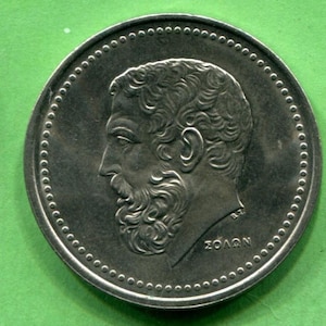 May include: A silver coin featuring a profile of a man with a beard and curly hair. The coin has a dotted border and text in Greek. The man's profile is facing left. The coin is on a green background.