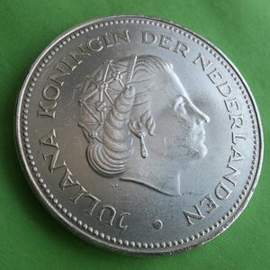 May include: A silver coin featuring a profile of a woman with an elaborate hairstyle and the text "JULIANA KONINGIN DER NEDERLANDEN" around the edge. The coin is set against a green background.