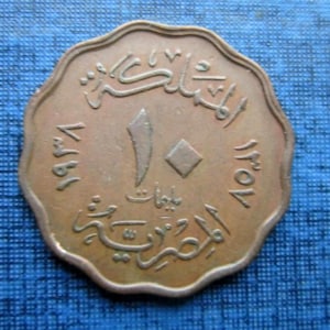 Puede incluir: Una moneda de color bronce con escritura y números árabes. La moneda tiene un borde festoneado y presenta un valor central de "10" con forma de diamante. El texto parece estar en árabe. La moneda está sobre un fondo azul.