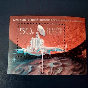 Può includere: Un francobollo sovietico vintage che presenta un'astronave su uno sfondo rosso e bianco. Il francobollo riporta la scritta "50k" e "ПОЧТА СССР 1989" in caratteri cirillici. Il francobollo è intitolato "МЕЖДУНАРОДНЫЙ КОСМИЧЕСКИЙ ПРОЕКТ "ФОБОС"" in caratteri cirillici.