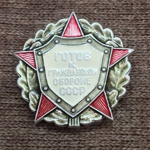 May include: A silver and red metal pin with a red star and a shield. The shield has the text "Готов к гражданской обороне СССР" in white.