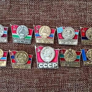 May include: Ten vintage enamel pins featuring the flags of the Soviet Socialist Republics. Each pin features a red flag with a hammer and sickle emblem and the name of the republic in Cyrillic script. The pins are in excellent condition and would be a great addition to any collection.