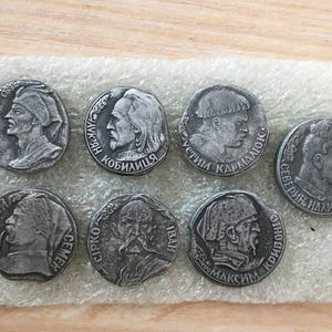 Könnte beinhalten: Sieben silberfarbene Metallmünzen mit Porträts historischer Persönlichkeiten und Text in kyrillischer Schrift. Die Münzen sind in einem Halbkreis auf einer hellbraunen Oberfläche angeordnet.