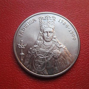 Puede incluir: Una moneda de plata con el retrato de una mujer coronada con joyas ornamentadas. La moneda tiene la inscripción "JADWIGA" y las fechas "1384-1399". La moneda está sobre un fondo rojo.