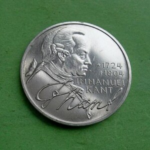 Puede incluir: Una moneda de plata con un perfil de Immanuel Kant, con los años 1724 y 1804. La moneda tiene la inscripción "IMMANUEL KANT" y una firma. La moneda está sobre un fondo verde.