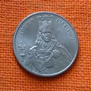 Puede incluir: Una moneda de plata con el retrato de una mujer coronada con joyas ornamentadas. La moneda tiene la inscripción "Ludwig" y las fechas "1784-1799". El fondo es de color naranja.