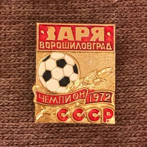 May include: A gold and red enamel pin featuring a soccer ball, a wheat stalk, and the text "ЗАРЯ ВОРОШИЛОВГРАД ЧЕМПИОН 1972 СССР".