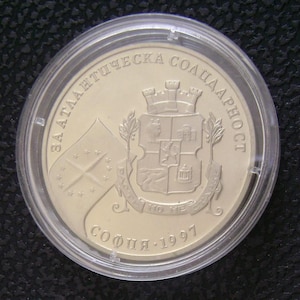 May include: A gold-colored coin encased in a clear plastic holder. The coin features text and a coat of arms with a flag design. The text includes the year 1997 and the city of Sofia. The coin is round.