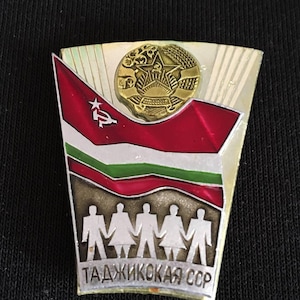 Könnte beinhalten: Eine silberne und rote Emaille-Anstecknadel mit einem goldenen Stern und einer roten Flagge mit Hammer und Sichel. Die Anstecknadel zeigt außerdem eine Gruppe von fünf Personen, die sich an den Händen halten, und den Text "ТАДЖИКСКАЯ ССР".