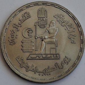 May include: A silver coin featuring an ancient Egyptian figure seated on a throne, holding a staff. The coin has Arabic script around the edge and a crescent moon symbol. The coin's design includes a brick-like structure and other symbols.