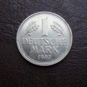 Könnte beinhalten: Eine silberne Deutsche Mark Münze von 1982. Die Münze zeigt die Zahl 1, die Worte "DEUTSCHE MARK" und das Jahr 1982. Zwei Eichenlaubzweige flankieren die Zahl 1. Die Münze liegt auf einer dunkelbraunen Lederoberfläche.