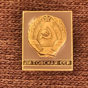 May include: A gold-colored pin with a red star and hammer and sickle emblem surrounded by a wreath of wheat. The pin has a light brown background and the text "ЛИТОВСКАЯ ССР" in black lettering.