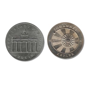 Peut inclure: Deux pièces de monnaie argentées. L'une représente la porte de Brandebourg avec le texte "HAUPTSTADT DER DDR BERLIN". L'autre pièce présente un motif de soleil rayonnant avec le texte "JOHANNES KEPLER" et les dates "1571" et "1630".