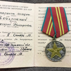May include: A close-up of a document with a red, yellow, and green ribbon medal. The document is titled "For Impeccable Service in the Ministry of Internal Affairs of the USSR." The document is dated October 8, 1958. The medal is a gold star with a hammer and sickle in the center.