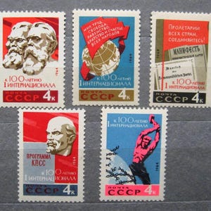 Op de afbeelding: Vier vintage postzegels van de Sovjet-Unie ter herdenking van de 100e verjaardag van de Eerste Internationale. De postzegels tonen afbeeldingen van Karl Marx en Friedrich Engels, een wereldbol, een hamer en een ketting, en een document met de tekst "PROLETARIERS VAN ALLE LANDEN, VERENIGT U!" De postzegels zijn rood, blauw en wit.