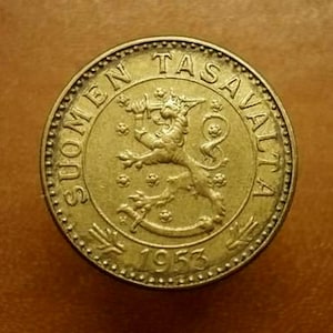 May include: A gold-colored coin featuring a lion rampant holding a sword, surrounded by text. The coin reads "SUOMEN TASAVALTA" and "1953". The coin has a detailed border and is set against a brown background.