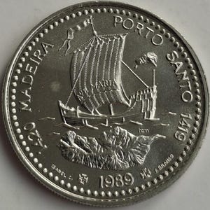 Puede incluir: Moneda de plata con una representaci&oacute;n detallada de un velero con la palabra "MARIA" en la vela, sobre un fondo de una isla monta&ntilde;osa. La moneda est&aacute; inscrita con "MADEIRA PORTO SANTO 1419" y el a&ntilde;o "1989".