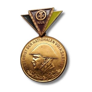 May include: A vintage gold-toned military medal. The medal features a profile of a soldier in a helmet and the text "FÜR DIENST DER NATIONALEN VOLKSARMEE". Above the medal is a triangular emblem with the letters "NVA".