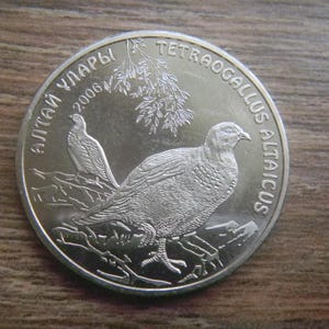 May include: A silver coin featuring two birds, one perched and one standing, with the text "TETRAOGALLUS ALTAICUS" and "2006". The coin has a detailed design of the birds and foliage.