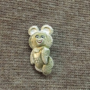 May include: A gold-colored pin featuring a cartoon bear with a smiling face. The bear is wearing a red shirt with a white circle and five smaller circles inside.