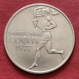 May include: A silver coin featuring a running figure holding a torch, with text in Cyrillic script. The coin reads "УНИВЕРСИАДА СОФИЯ 1977". The design is a relief, with the figure and text raised against a smooth background. The coin has a circular shape.