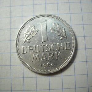 Op de afbeelding: Een zilveren 1 Deutsche Mark munt uit 1963. De munt toont het nummer 1, de woorden "DEUTSCHE MARK" en het jaar 1963. Eikenbladeren staan aan weerszijden van het nummer 1. De munt ligt op een rasterachtergrond.