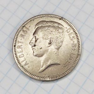 May include: A silver coin with a profile of a man, the text "ALBERT ROI" and "DES BELGES" around the edge. The coin has a detailed design and a textured border, set against a grid background.