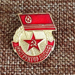 May include: A red and gold metal pin with a red star and hammer and sickle, surrounded by a wreath of wheat. The pin has the text "Инструктор ДОСААФ" in red lettering.