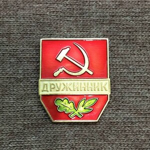 May include: A red and gold enamel pin with a hammer and sickle and the word "DRUZHINNIK" in Cyrillic script. The pin also features a green oak leaf.